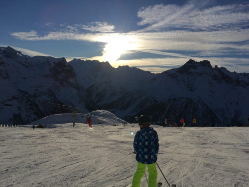 Au premier plan, on voit mon fils de dos sur ses skis prêt à descendre une piste, en second plan, la crête des montagnes et en arrière plan le ciel bleu parsemé de nuages et un soleil en train de se coucher
