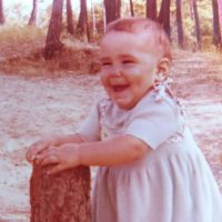 Sophie, bébé joufflu à un an dans une robe avec un sourire aux lèvres. Les mains sur un tronc d'arbre pour se tenir en équilibre. La photo est un peu passée en couleurs.