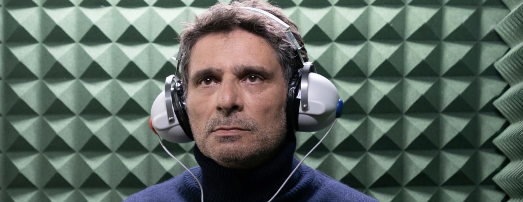 Pascal Elbé est dans une cabine d'audioprothésiste. Il a un casque sur les oreilles et a un air concentré.