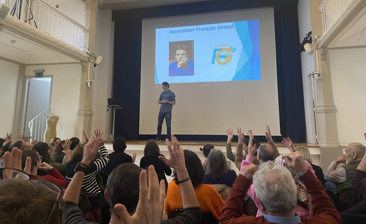 Slide projetée à l'écran avec la photo de François à gauche et le logo de l'association à droite (AFG), David Rousseff, ancien président de l'association, ouvre la séance de l'assemblée générale avec en premier plan les membres qui applaudissent en levant les bras et secouant les mains pour dire bravo.