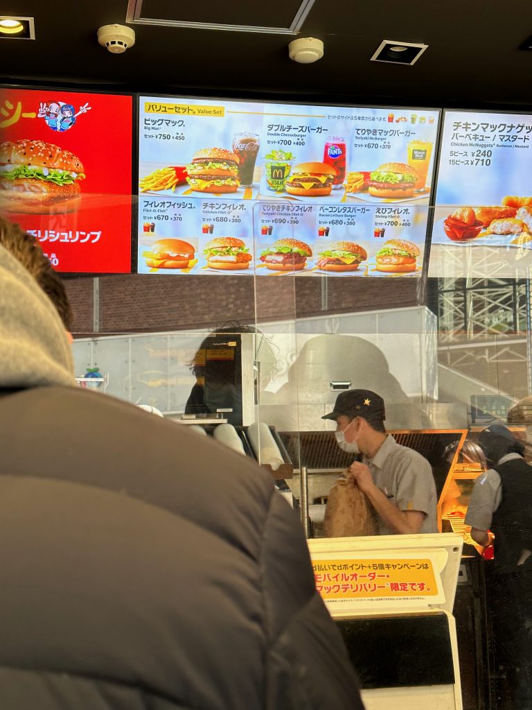 Panneaux de menus du macDonalds en japonais. On voit le visuel des hamburgers et des menus écrits en japonais doublés de l'anglais, avec le prix en yen qui va de 670 yens à 750 yens pour le menu. Au premier plan, il y a une caisse électronique avec une inscription en caractères japonais sur un panneau jaune.