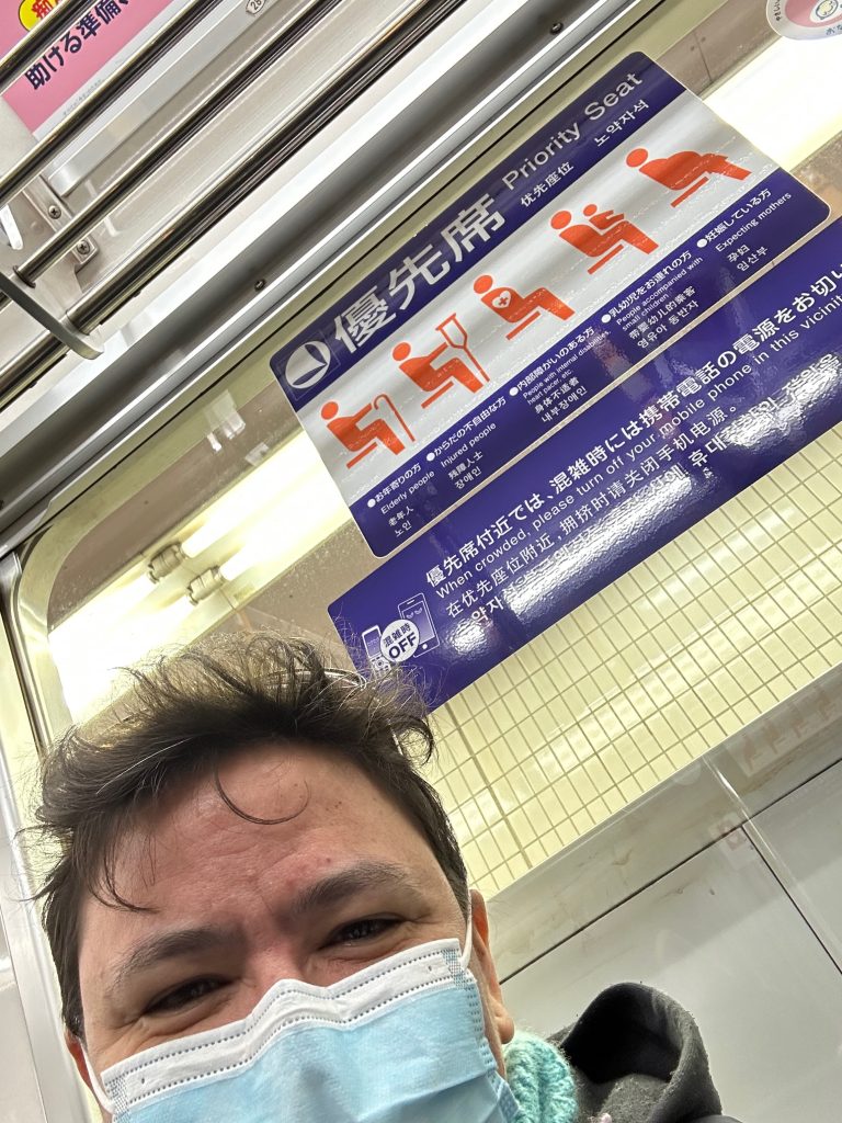 Photo de l'autocollant placé au dessus des places prioritaires qui sont réservés aux personnes agées, personnes blessées, personnes avec un handicap invisible (pacemaker, etc…), personnes accompagnées de petits enfants et femmes enceintes. Je suis au premier plan masquée, on ne me voit que les yeux sourieurs et les cheveux un peu désordonnés. Il est indiqué plus bas d'éteindre son téléphone portable ou de le mettre en silencieux.