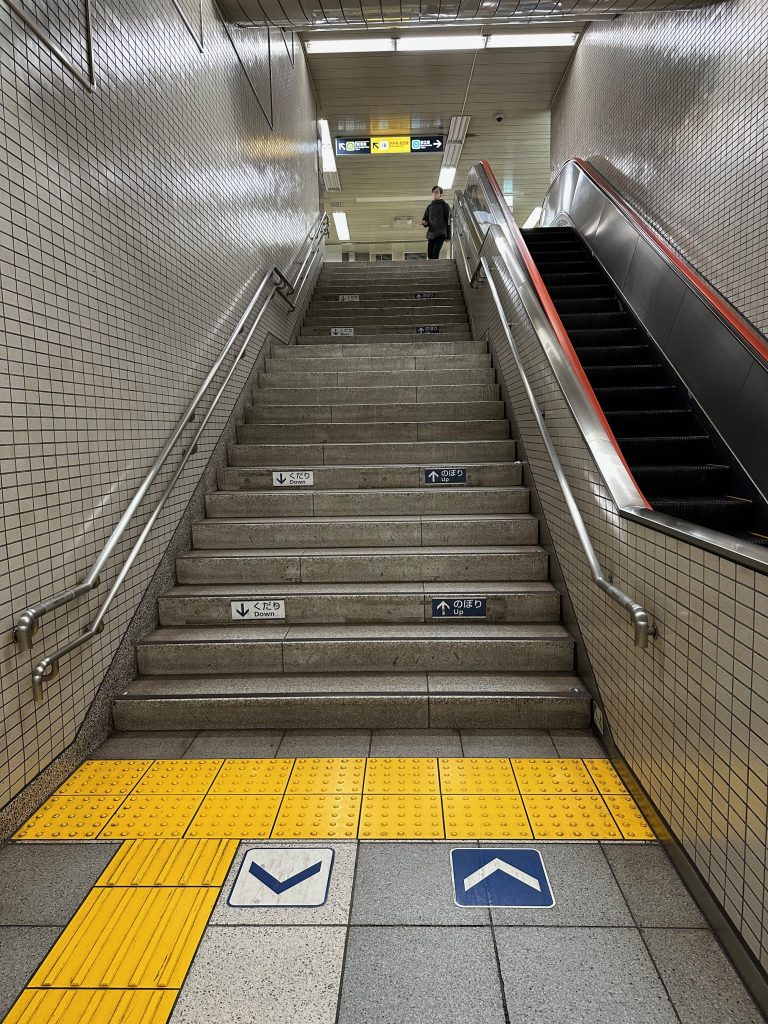 escalier avec bande podotactile à picots au début de l'escalier et une bande verticale sur la gauche de l'image. Deux flèches sont indiquées à plusieurs reprises, celle qui sert à descendre est sur la gauche, celle qui sert à monter est sur la droite. Sur la droite de la photo, on a un escalator qui monte.