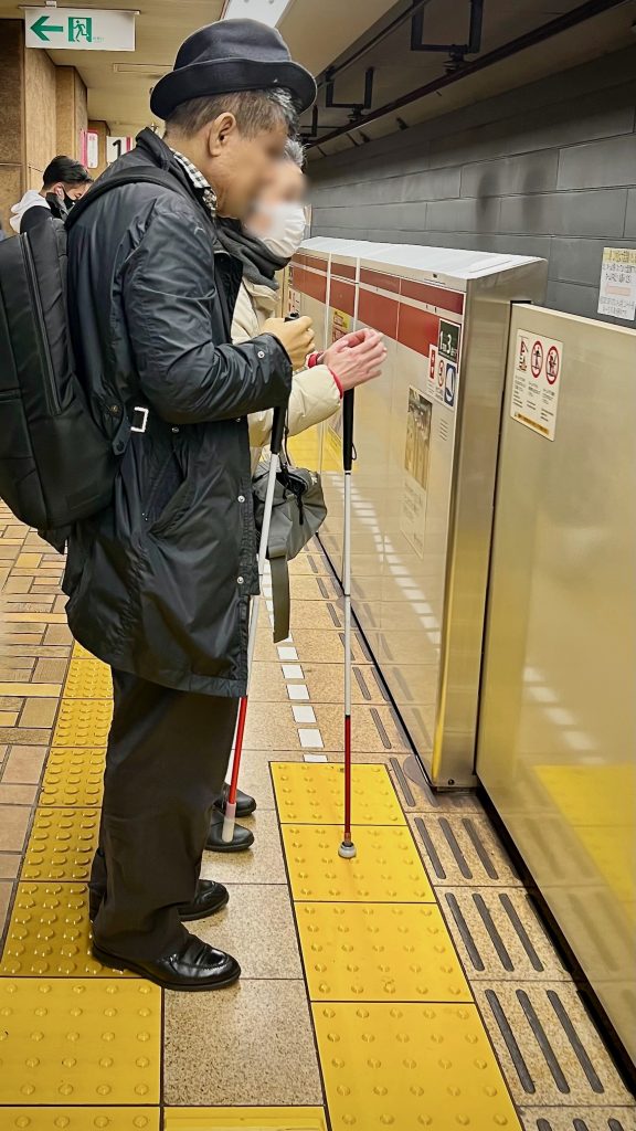 Un homme et une femme attendent le métro à un emplacement où il y a des places prioritaires dans le wagon. Ils sont placés sur la bande podotactile à picots. Ils sont aveugles tous les deux et ont tous les deux une canne, face à une porte. Leur visage est flouté par respect de la vie privée.