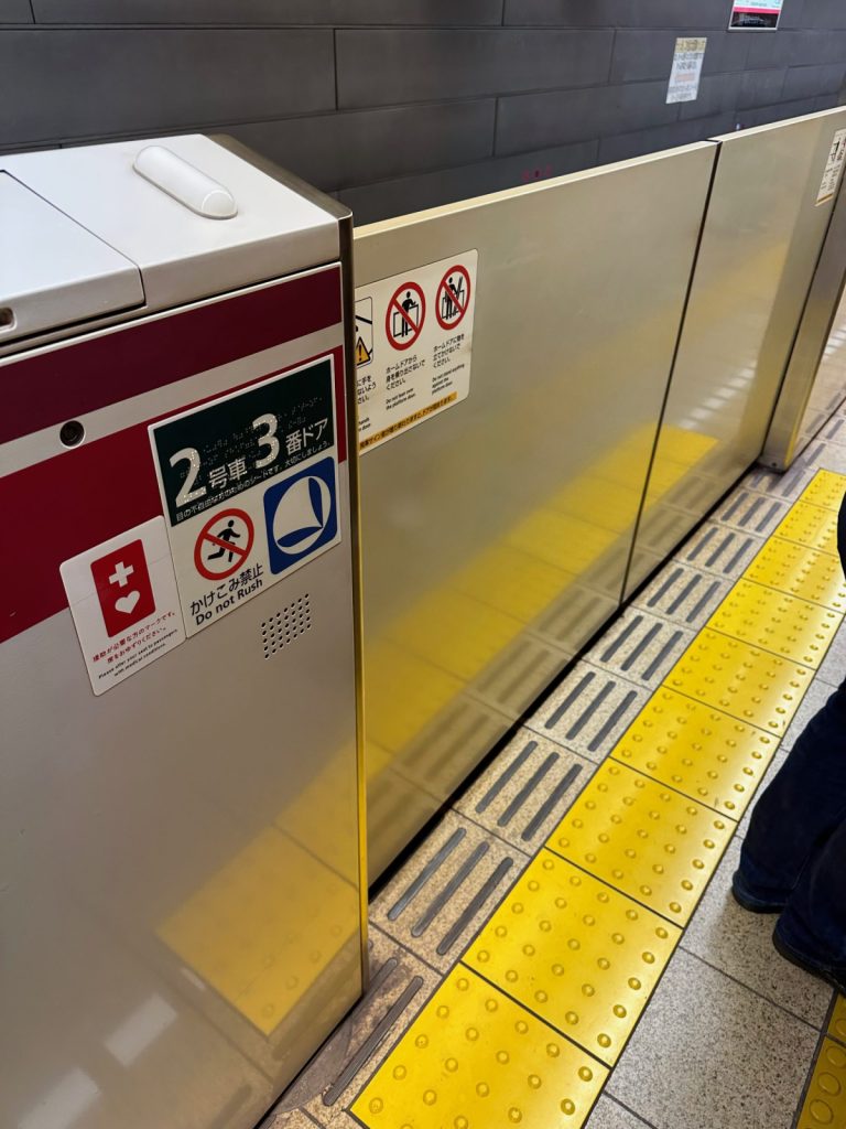 Gros plan de la porte de métro avec au bord du quai une bande verticale suivie d'une bande à pictos de couleur jaune vif. Sur la séparation métallique, on aperçoit le numéro du wagon de la rame ainsi que le numéro de la porte. Il est indiqué qu'il ne faut pas courir et qu'il y a des places prioritaires à cette porte par le biais d'un logo bleu qui symbolise le profil d'une chaise. Sur le dessus de cette séparation métallique, on y voit une bande lumineuse blanche pour les personnes sourdes. Un pictogramme de croix et de cœur est également présent dans un rectangle rouge (défibrillateur je pense) et enfin, les pictogrammes à destination du grand public (ne pas enjamber la protection, ou de s'y accouder)