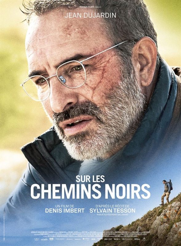 Affiche du film "sur les chemins noirs". Jean Dujardin avec une paire de lunettes cerclées, le regard dans le vide en arrière plan avec au premier plan une autre vue de l'acteur qui descend la montagne. Un film de denis imbert d'après le récit de sylvain tesson publié aux éditions gallimard.