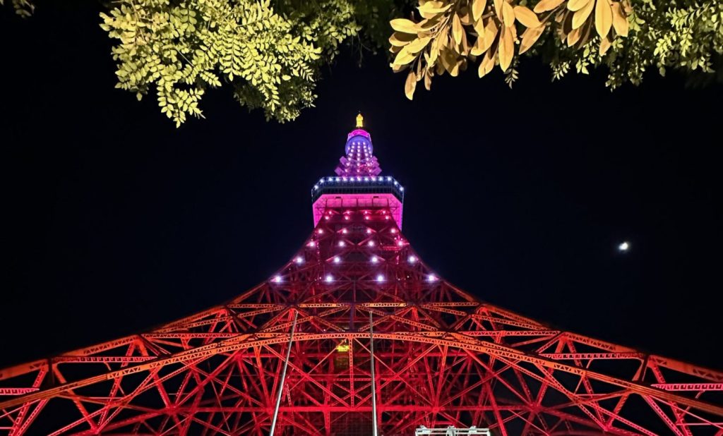 Tour de Toyko vue d'en bas, les couleurs vont du rouge au violet qui est au sommet de la tour. Il fait nuit, on voit le feuillage des arbres sous lesquel j'étais pour prendre la photo.