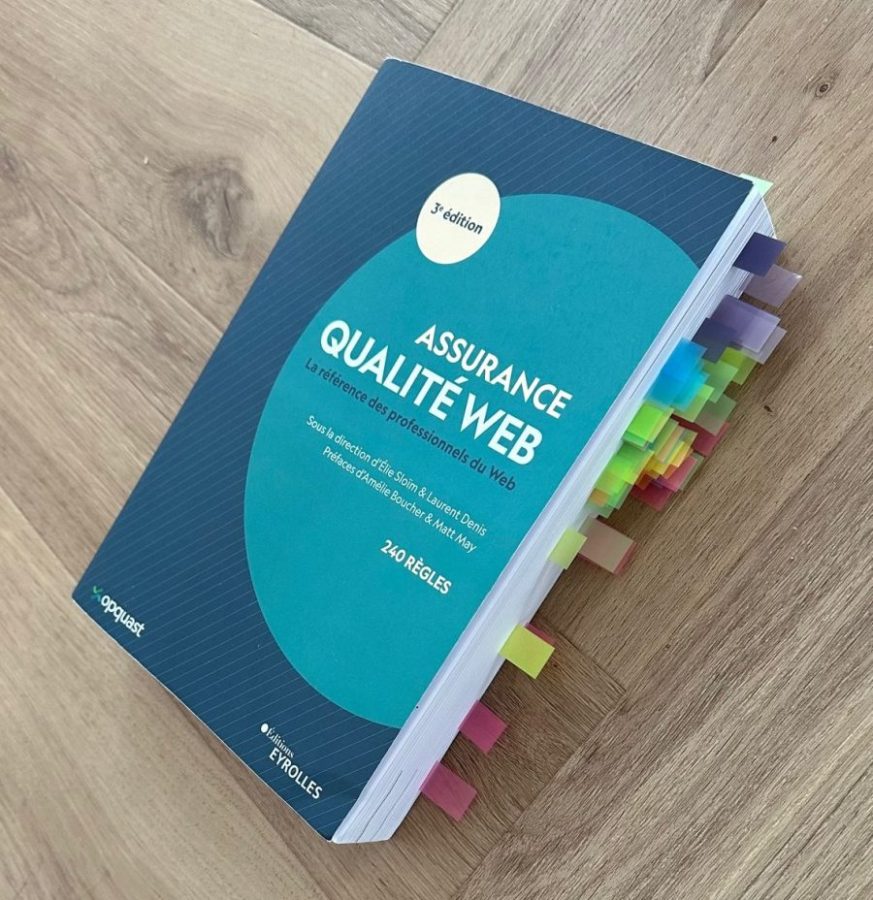 Livre avec plein d'onglets collés sur les rebords des pages, posé sur un sol en bois. La couverture indique : Assurance Qualité Web. La référence des professionnels du web, sous la direction d'élie sloïm et laurent denis. Préfaces d'amélie boucher et matt may. 240 règles.
