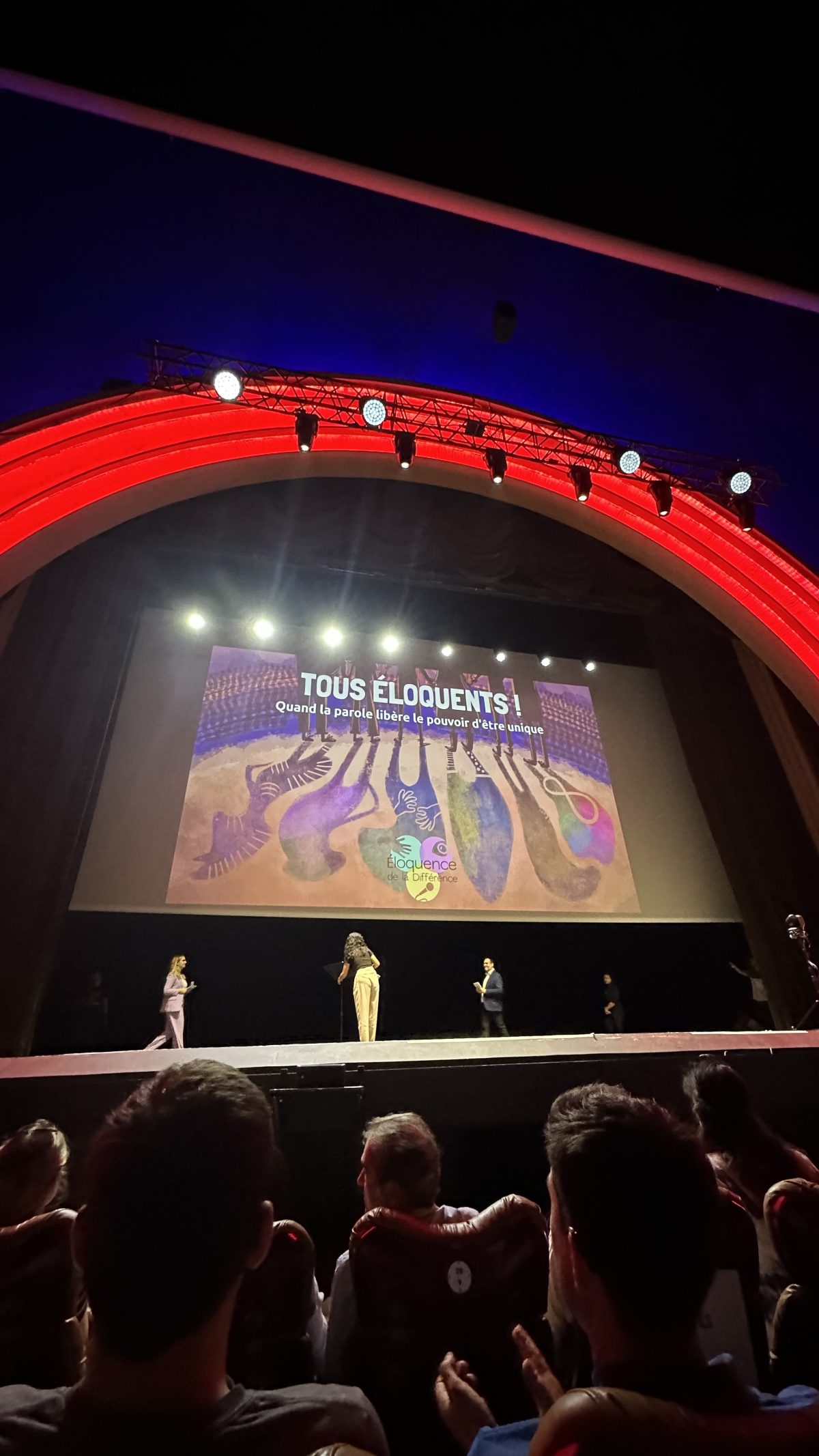Prise de vue de la scène du grand rex, le demi-cercle rouge si emblématique, avec une projection de l’affiche de Tous éloquents ! Il est écrit sur cette affiche « Quand le parole libère le pouvoir d’être unique »